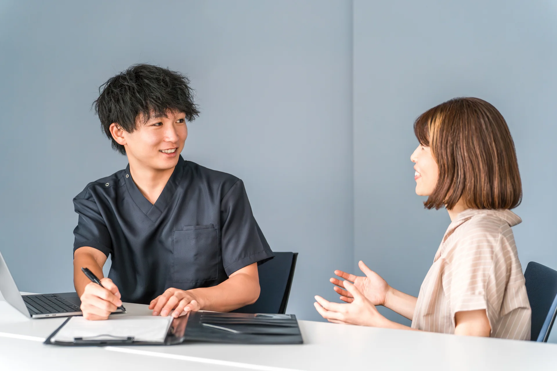 スクラブ姿の男性にメモを取ってもらいながら相談する女性（診察で頬の悩みを伝えるイメージ）