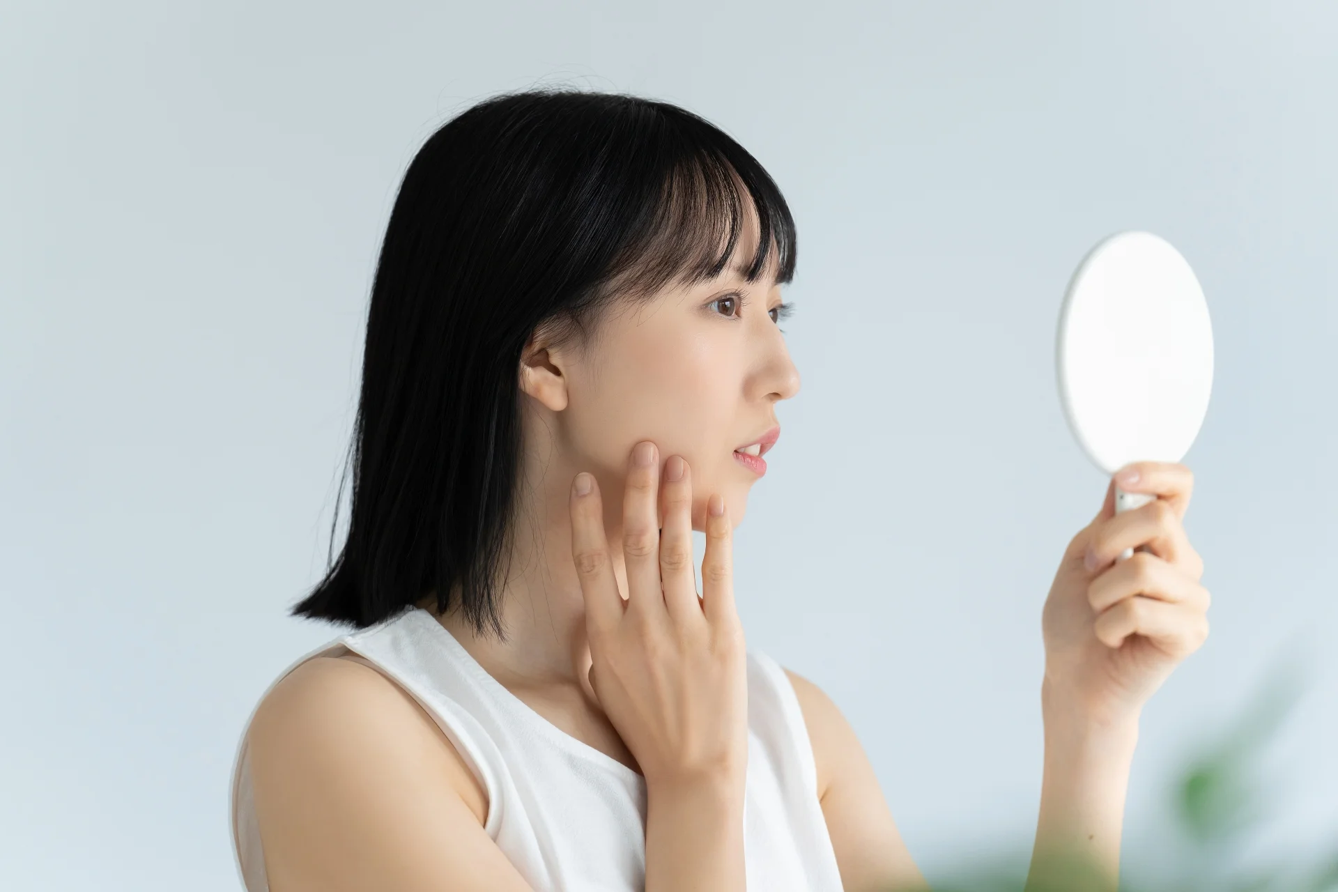 手鏡を持ち、あごに指を添えて顔を確かめる女性(将来の顔の変化についての疑問のイメージ)