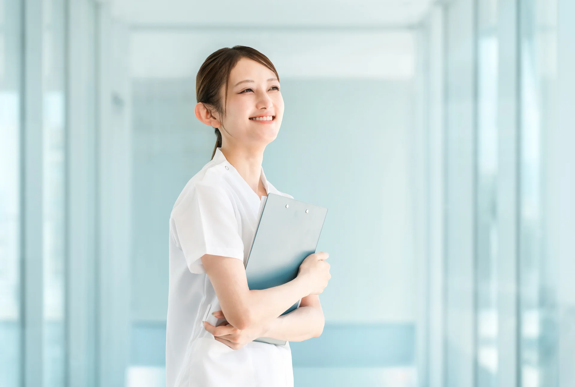 白衣の女性スタッフがクリップボードを抱えてほほえむ様子(フェイスライン整形の疑問を相談するイメージ)
