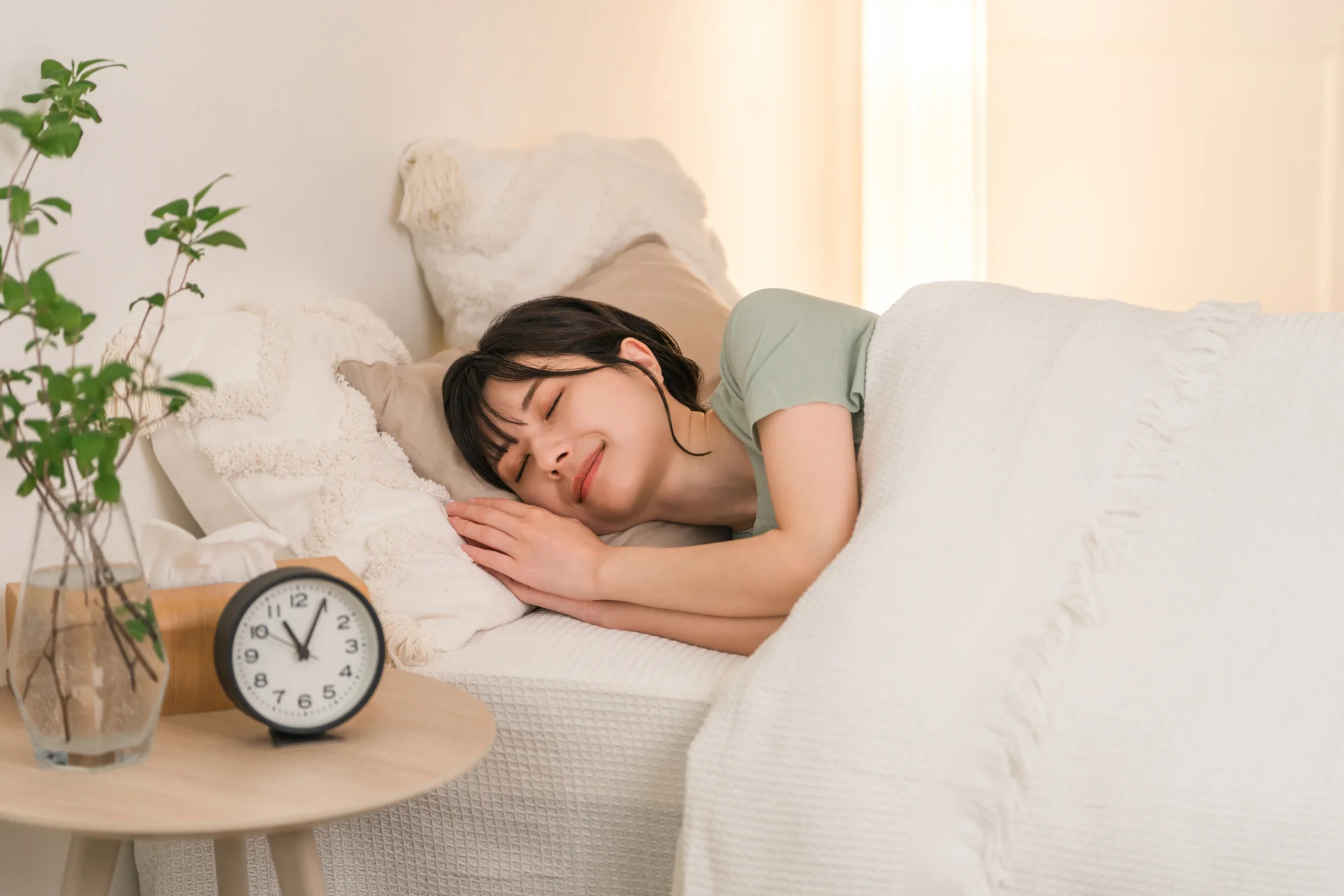 目覚まし時計を置いたベッドで眠る女性。睡眠をとって翌朝のむくみを減らすイメージ