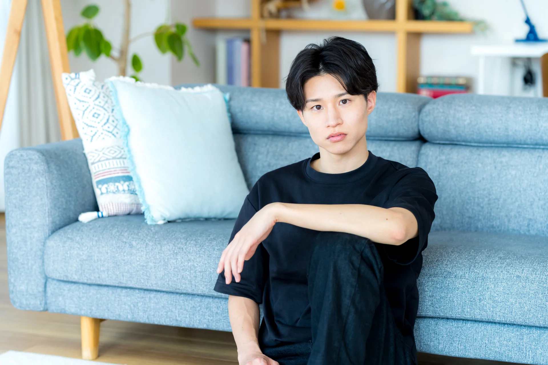 ソファの前の床に座る黒い服の若い男性（頬肉の疑問を考えるイメージ）