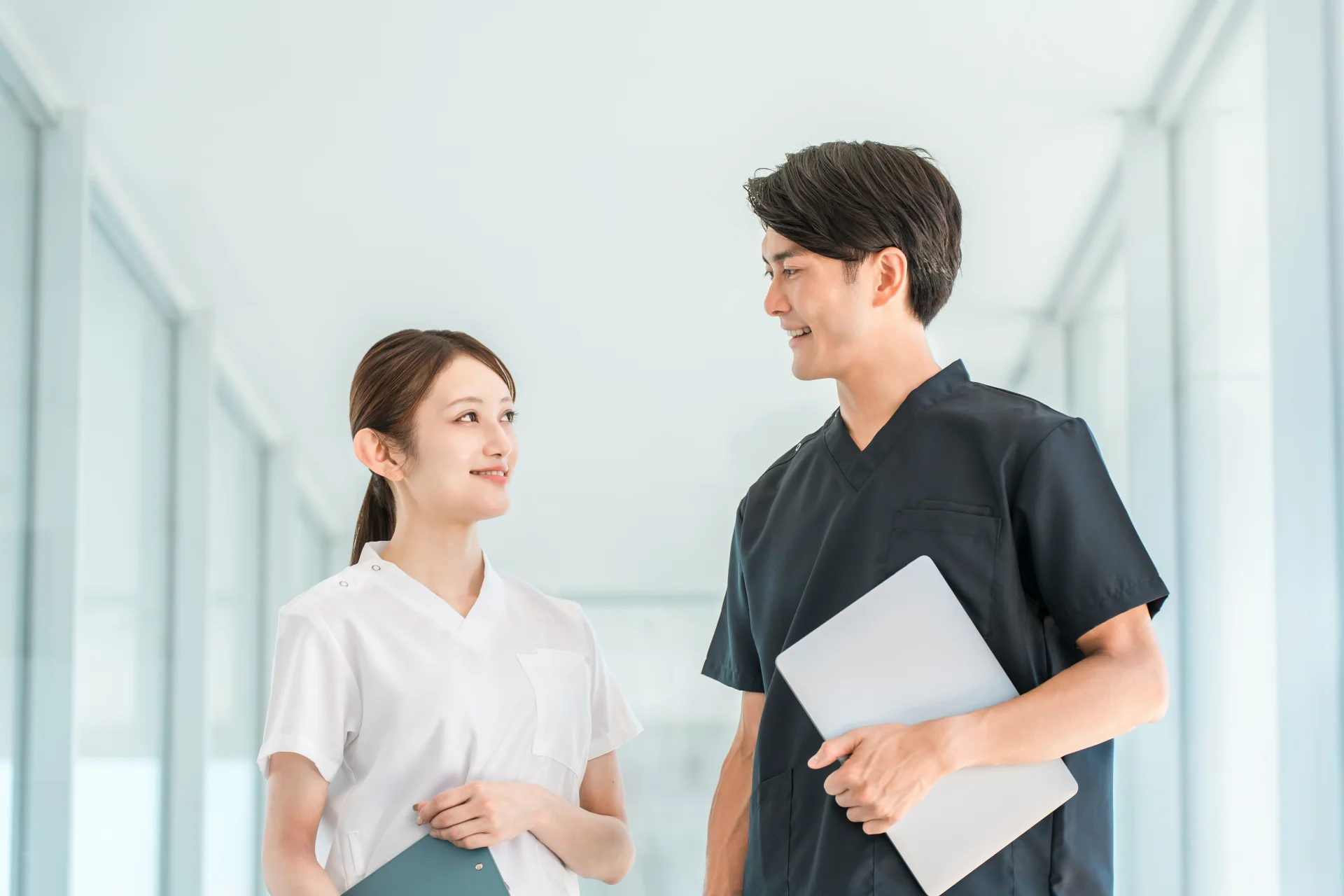 白いスクラブの女性スタッフと紺のスクラブで端末を持つ男性スタッフが院内の廊下で話す様子(小顔整形の疑問を医療スタッフに聞くイメージ)