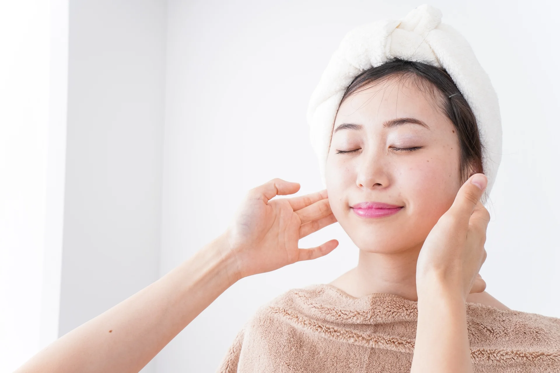 頭にタオルを巻いた女性が目を閉じて両手で耳の下から頬に触れている様子(自分の顔の輪郭を確かめて小顔整形の候補を考えるイメージ)