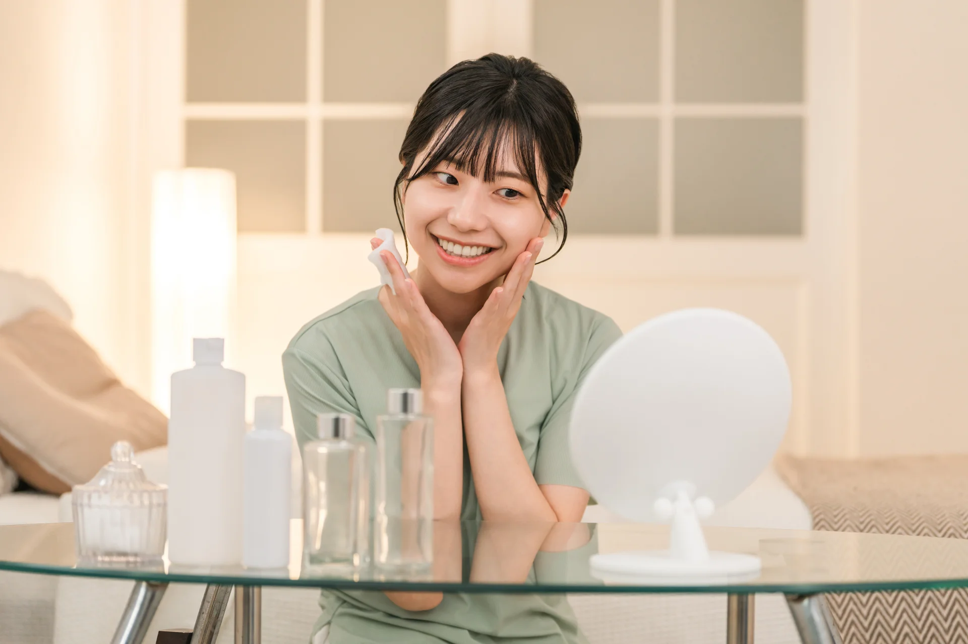 鏡を置いたテーブルの前でコットンを持ち両手を頬に添えてほほえむ女性